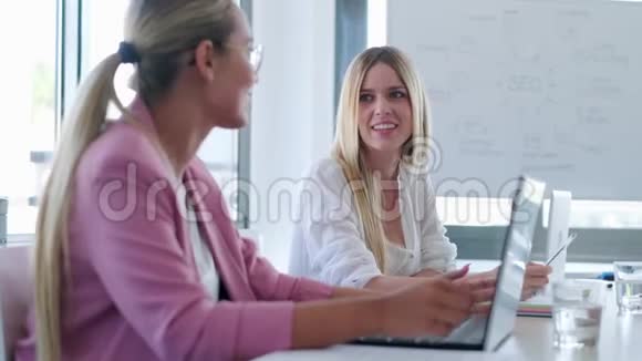 两个微笑的年轻女商人在同事的空间里用笔记本电脑聊天视频的预览图