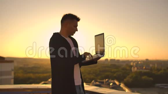 镜头跟随着一个人在日落时分带着笔记本电脑和一个瓶子穿过屋顶自由职业者打开笔记本电脑视频的预览图