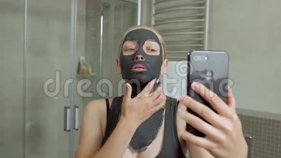 一个女人在镜子附近的浴室里脸上戴着一个黑色的泥面罩在智能手机摄像头上拍摄自己的照片视频的预览图