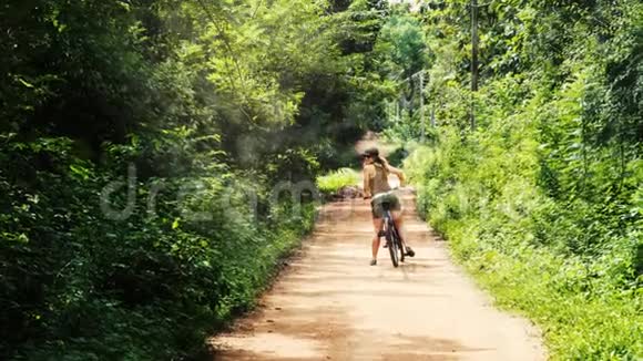 一个女孩戴着帽子和太阳镜骑自行车看着她的肩膀的视差动画在斯里兰卡的土路上视频的预览图