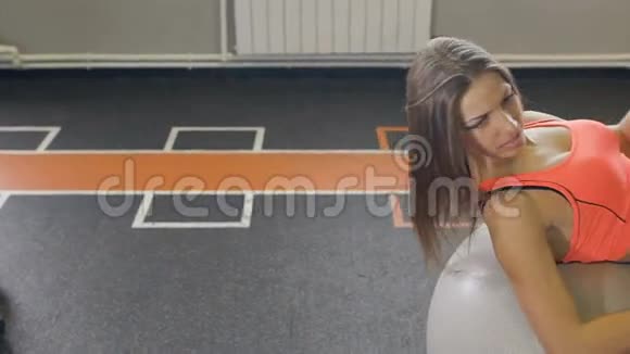健身房里的健身女人靠普拉提球休息年轻女子在健身球上做运动年轻女孩的粉色健身视频的预览图