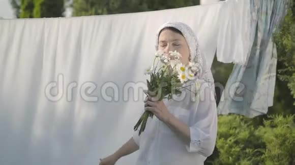 漂亮的高级妇女头上戴着白色披肩闻着雏菊看着相机微笑着看着晾衣绳视频的预览图