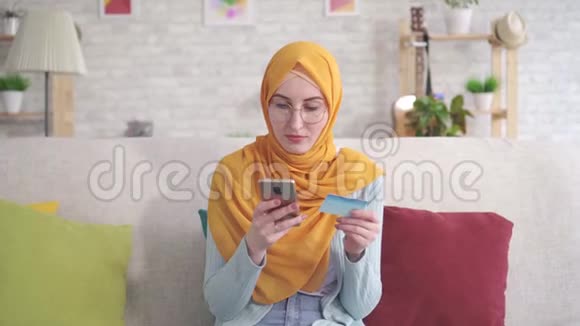 戴着头巾拿着智能手机坐在客厅沙发上的银行卡的年轻穆斯林妇女的不幸和烦恼的肖像视频的预览图