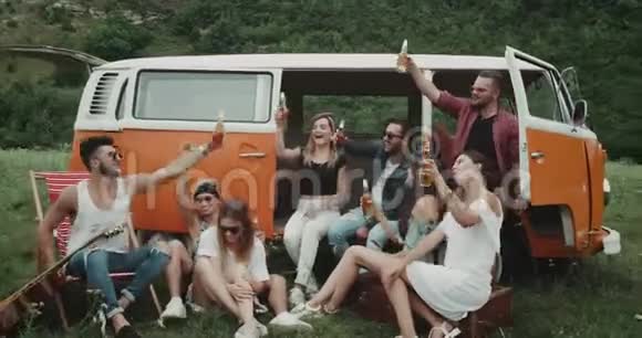 一大群朋友在野餐聚会上喝着啤酒和欢呼声坐在一辆复古面包车后面视频的预览图
