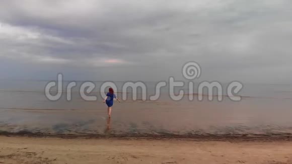 空中年轻女子穿着蓝色鲜艳的裙子跑步浅水风景波罗的海的黄色日落景观视频的预览图