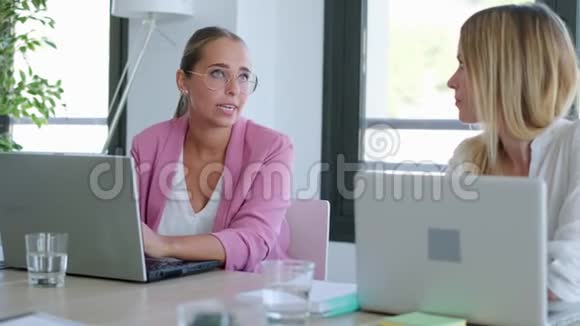 两个微笑的年轻女商人在同事的空间里用笔记本电脑聊天视频的预览图