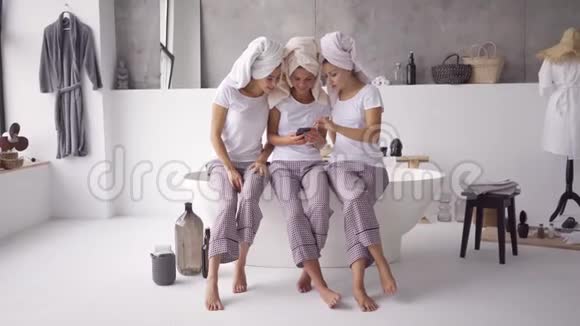 三个快乐的女朋友穿着同样的睡衣头上有毛巾在一起看手机视频的预览图