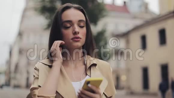 戴着耳机的美丽少女喜欢用智能手机听音乐和微笑在城市散步视频的预览图