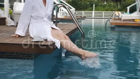 优雅的女人穿着白色浴袍坐在豪华别墅的泳池边的码头上在泳池里着脚视频的预览图