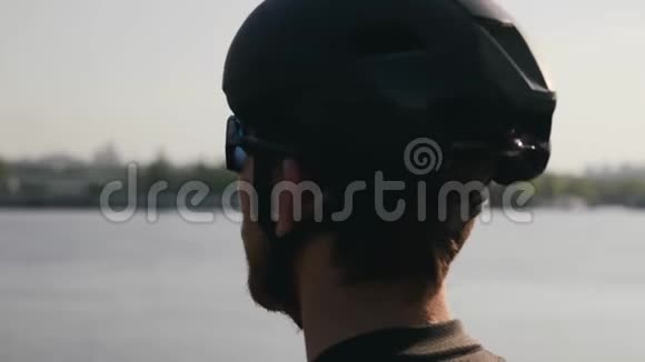 戴着黑色头盔和太阳镜的有胡子的骑自行车的人目光集中到地平线上河和桥梁的背景骑自行车视频的预览图
