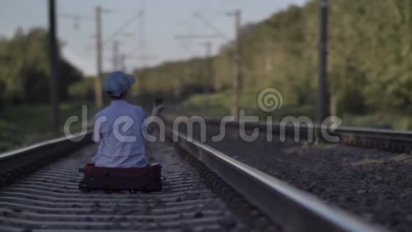 戴着帽子的悲伤男孩坐在户外铁路上的手提箱上视频的预览图