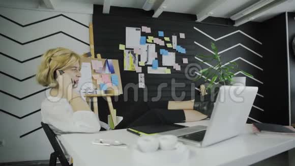 在现代的办公室里女人用电话说话双腿坐在桌子上视频的预览图