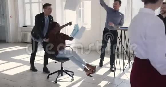 快乐迷人的黑人商务女性庆祝成就与同事骑着椅子和扔纸在空中视频的预览图