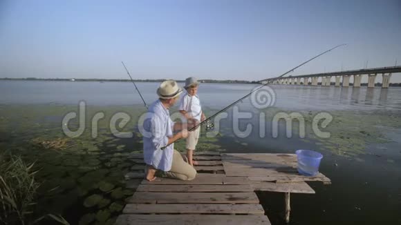 快乐的父亲和小儿子在河边的码头上戴着草帽暑假期间在村里钓鱼视频的预览图