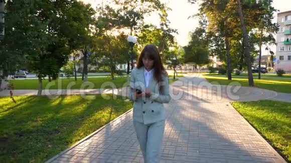 穿浅色西装的女人手里拿着平板电脑上班美丽的女商人沿着公园的小巷散步视频的预览图