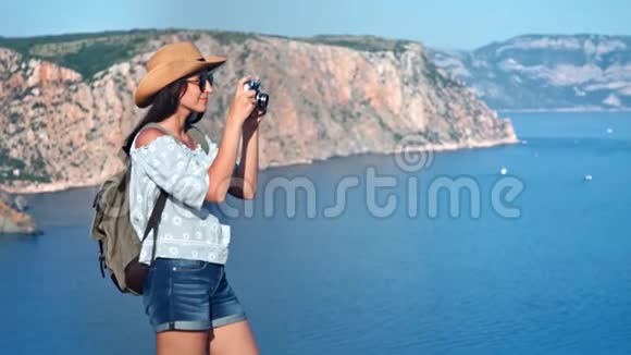 迷人的旅行女性戴着帽子和太阳镜用相机拍摄海景照片视频的预览图