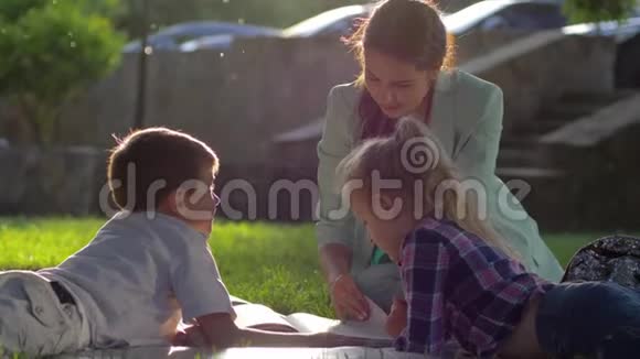 学前教育美丽的女性为小男孩和女孩在阳光明媚的户外草地上坐着看书视频的预览图
