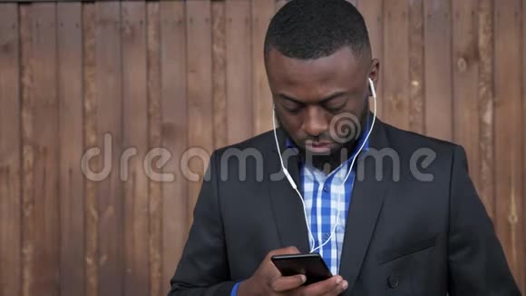 戴着耳机的黑人正在黑木背景上浏览手机视频的预览图
