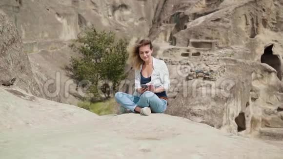 一个穿着蓝色牛仔裤的金发女郎坐在石头上看着电话古城的背景视频的预览图