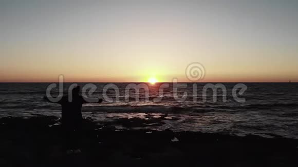 年轻女性的剪影站在多石的海滩上双手在夕阳下的波浪海上伸展开来视频的预览图