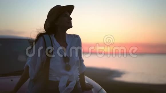 性感的旅行女人坐在车盖上在猩红的日落海景背景全景视频的预览图