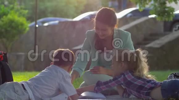 快乐的童年微笑的妈妈在阳光下为坐在绿色草地上的小男孩和小女孩读书视频的预览图