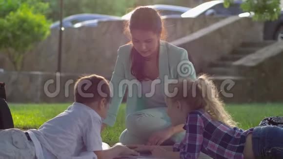 在户外看书快乐的女家庭教师为小男孩和小女孩在阳光明媚的露天草地上坐着看书视频的预览图