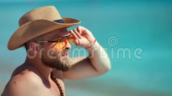 英俊的欧洲男人戴着太阳镜和帽子在海滩上放松海景背景视频的预览图