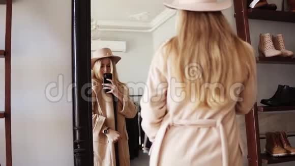 漂亮的金发女孩站在镜子前穿着米色外套和时髦的帽子和她一起拍了一张照片视频的预览图