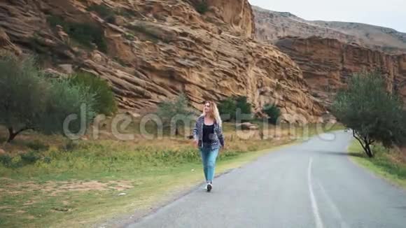 一个微笑的年轻金发女人穿着牛仔裤和毛衣的简单着牛仔裤和毛衣走在路上的背景视频的预览图