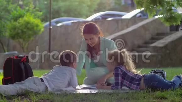 开朗的女教育家为小男孩和小女孩在阳光明媚的阳光下坐在大自然的绿草上读书视频的预览图