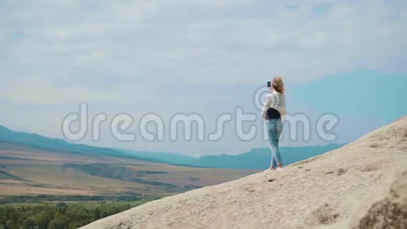 一个金发女孩站在一座山的顶上背景是湛蓝的天空她用手机举起了手视频的预览图