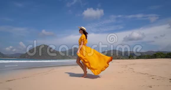 穿着黄色连衣裙戴着草帽的年轻美女模特在海边漫步白沙蓝天和视频的预览图