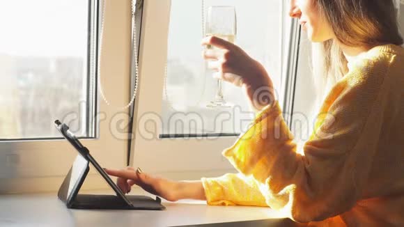 穿黄色外套的年轻女孩喝香槟在平板电脑上看新闻视频的预览图