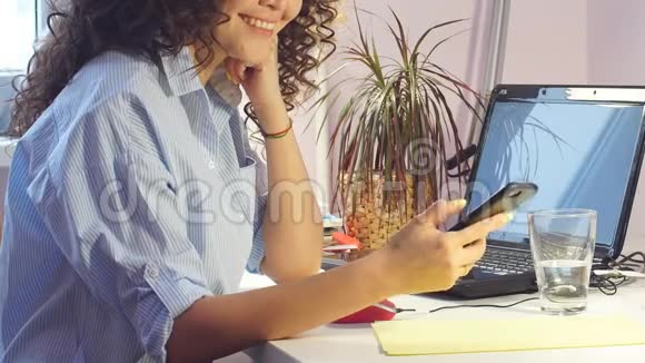年轻的微笑的女人在家里的办公室里坐在电脑前用智能手机视频的预览图