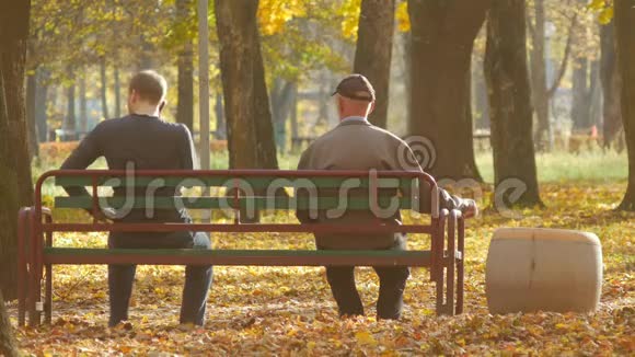 秋天阳光明媚的一天一个年轻人和一个老人坐在公园的长凳上不同年龄的一代视频的预览图
