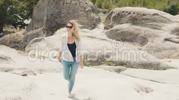 一位年轻的金发女郎穿着牛仔裤白色衬衫和太阳镜走在被刻在岩石上的古城附近视频的预览图