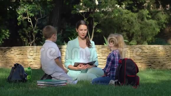 年轻的女教师在户外为小男孩和小女孩上课手里拿着书坐在草地上视频的预览图