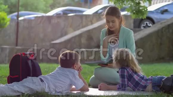 放学后小学童在阳光明媚的阳光下在大自然中坐在绿色草坪上听着女孩子读有趣的书视频的预览图