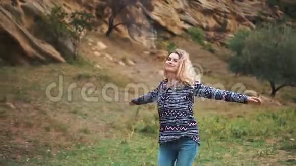 漂亮的年轻漂亮的女人金发飞扬在绿色的田野和陡峭的悬崖的背景下转过身视频的预览图