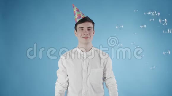 戴着派对帽的年轻人惊讶地在蓝色背景上飞泡泡视频的预览图