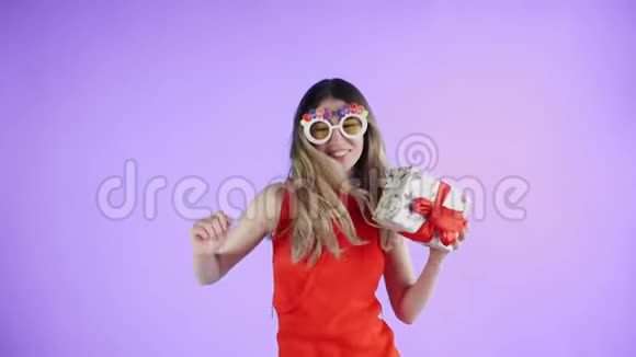 年轻女子戴着滑稽的眼镜手里拿着礼物在紫色的背景上跳舞视频的预览图
