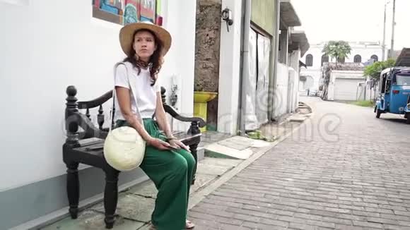 戴着草帽的漂亮时髦女人坐在街边的长凳上视频的预览图