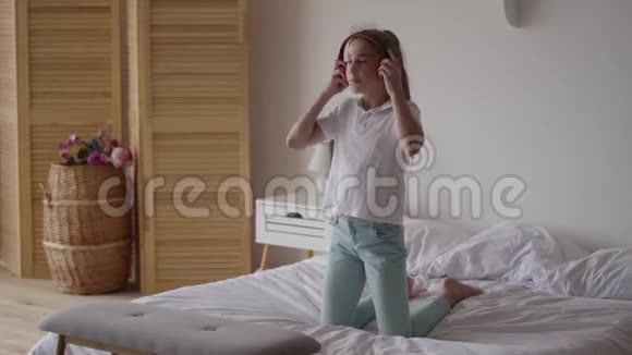 有趣可爱的小女孩戴上耳机试图跳舞摔倒在床上年轻的音乐爱好者玩得开心视频的预览图