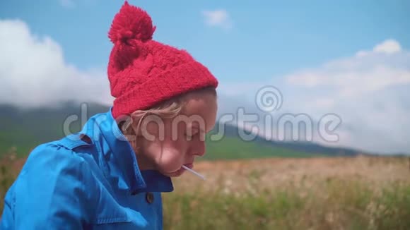 有趣的女孩站在阳光明媚的夏天金发女郎穿着蓝色外套戴着红色帽子嘴里放着三个棒棒糖玩得很开心视频的预览图