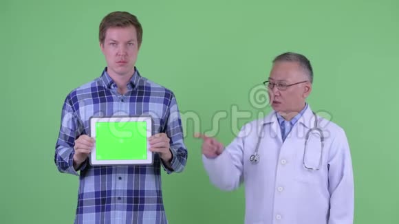 穿着成熟的日本男医生年轻男子展示数码平板电脑并竖起大拇指视频的预览图
