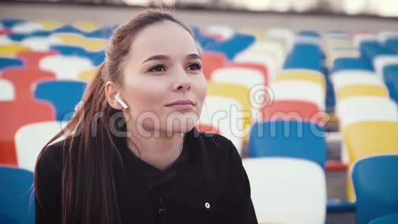 一个女孩坐在运动场的观众排看着训练通过耳机听音乐视频的预览图