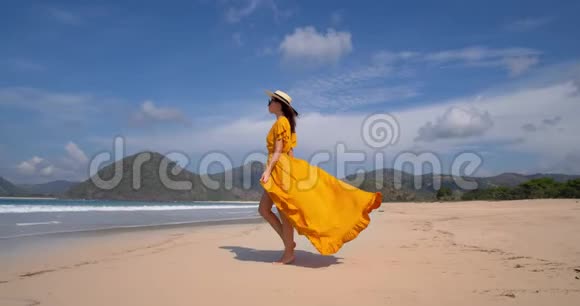 穿着黄色连衣裙戴着草帽的年轻美女模特在海边漫步白沙蓝天和视频的预览图