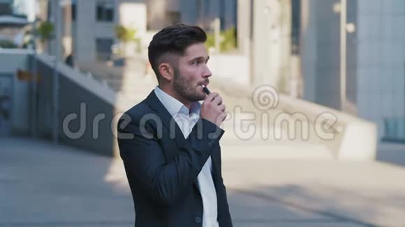 年轻英俊的商人在西装使用智能手机和吸烟IQOS附近的大办公楼商业生活方式视频的预览图