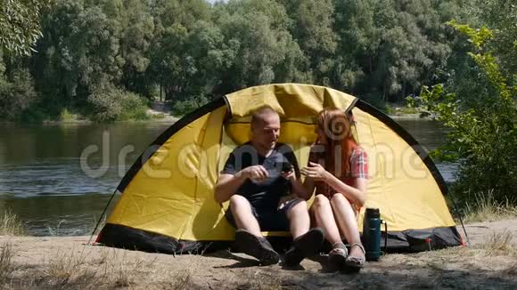 心满意足的幸福夫妇坐在帐篷里旅游和旅游的概念女孩用热水瓶倒茶森林和河流视频的预览图